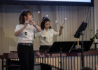 Eine Frau in schwarz-weißer Uniform spielt stehend Querflöte. Im Hintergrund sieht man eine weitere Frau, die mit vier Schlägern Marimba spielt. Am rechten Bildrand erkennt man eine Klarinettenspielerin.